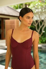 A woman wearing a structured burgundy one-piece swimsuit with thin shoulder straps, standing in front of a tropical poolside background.