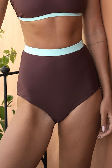 A woman modeling a two-piece bikini set in a dark brownish-purple color with light blue trim, standing against a peach-colored wall next to a potted plant.