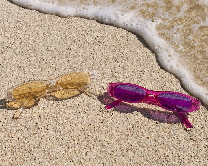 Two pairs of sunglasses, one gold and one pink, on a sandy beach with ocean waves.